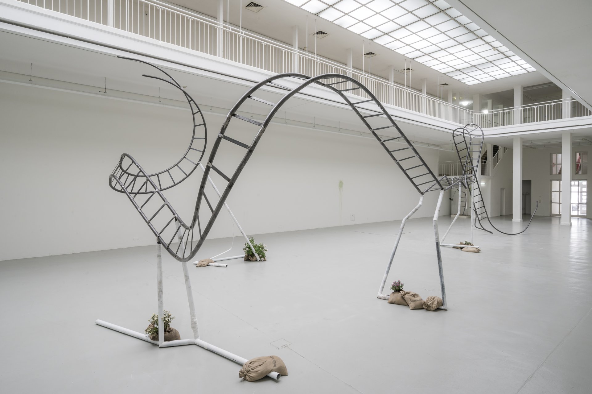 Une grande sculpture métallique ressemblant à un rail de montagnes russes tordues se dresse dans une galerie blanche et spacieuse dotée d'un haut plafond et de puits de lumière ; de petites plantes et des sacs de sable sont placés à sa base.