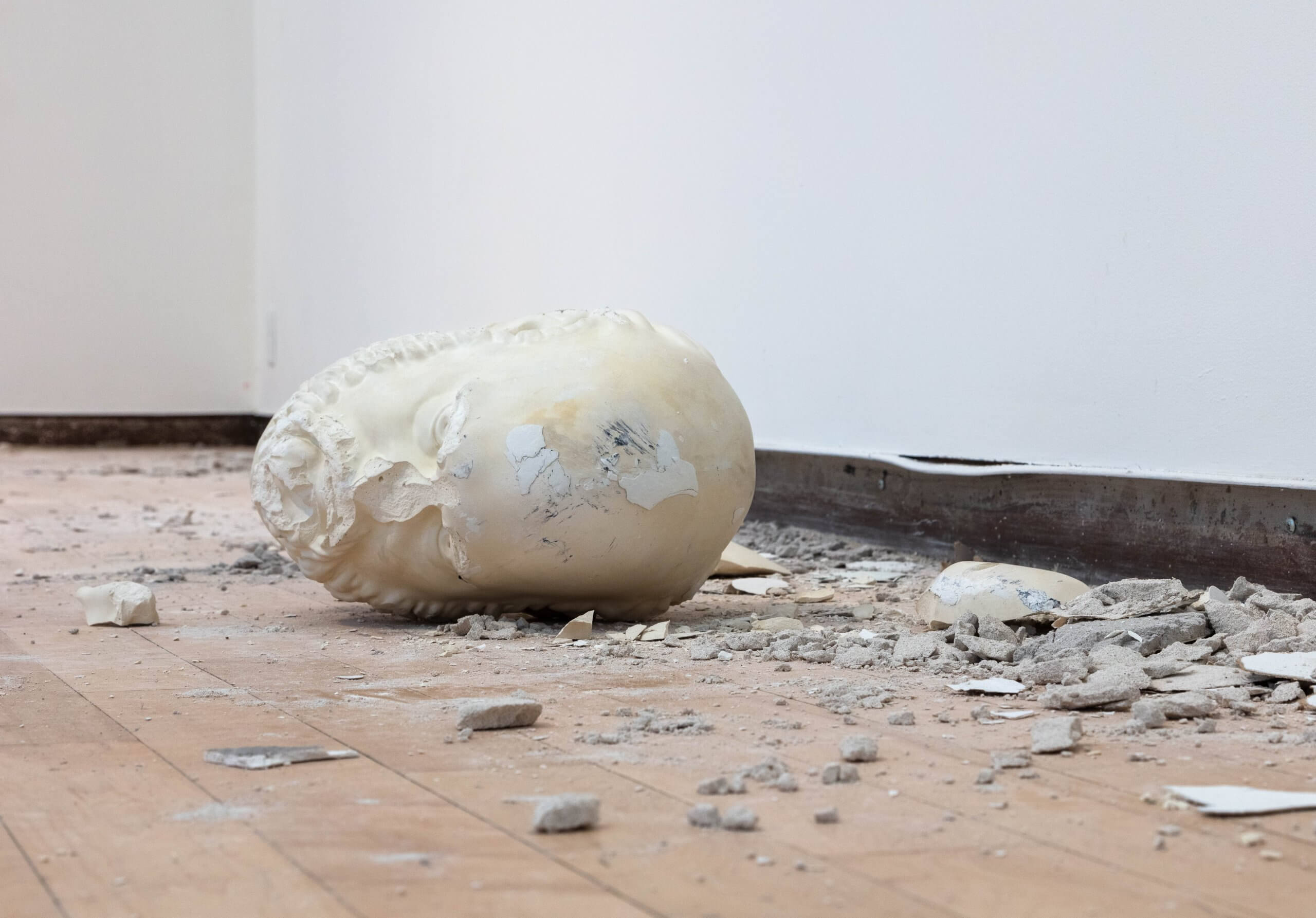 Une tête en plâtre brisé repose sur un sol en bois, entouré de débris et de fragments de plâtre épars, près d'un mur blanc.