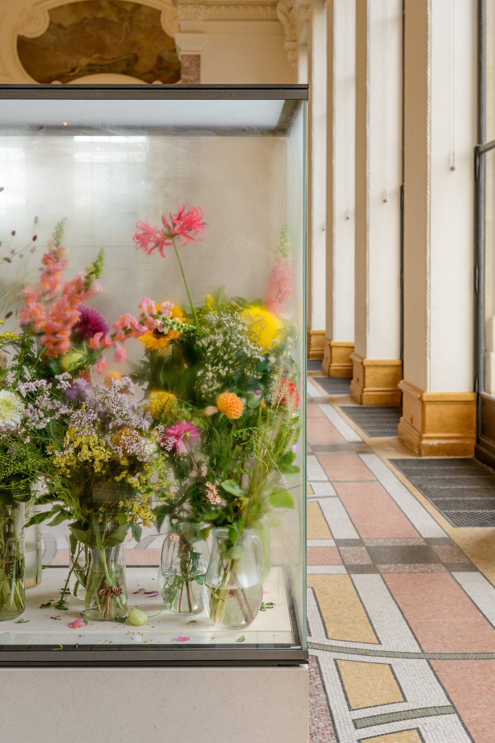 Une vitrine remplie de fleurs colorées assorties trône dans une pièce lumineuse dotée de grandes fenêtres et d'un carrelage à motifs.