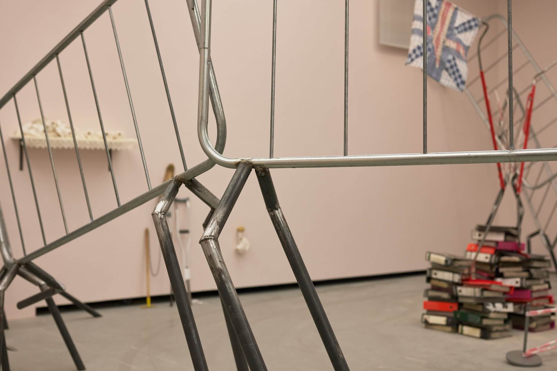 Gros plan sur des barrières métalliques pliées dans une pièce où se trouve une installation artistique, avec une pile de livres et d'autres objets à l'arrière-plan.