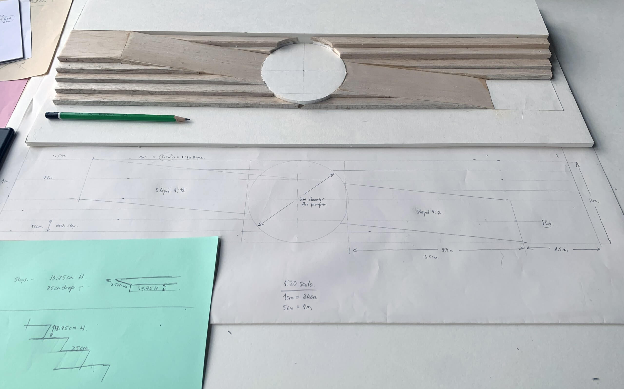 Un espace de travail avec un modèle en bois, un crayon, une note verte avec un croquis et un grand dessin technique avec des mesures et des détails de construction.