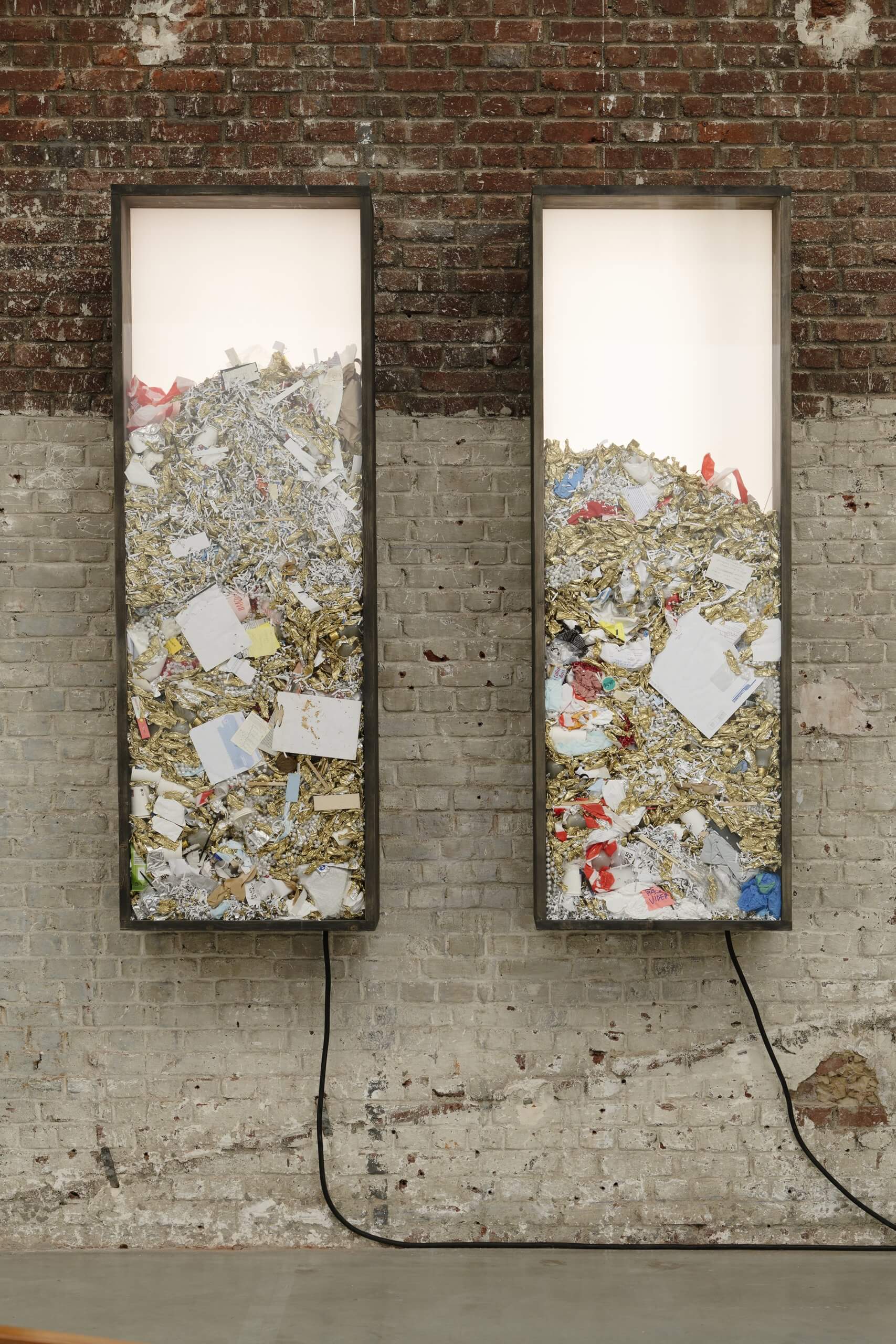 Deux vitrines verticales montées sur un mur de briques, chacune remplie de divers déchets et papiers, avec des cordons électriques partant du bas de chaque vitrine.