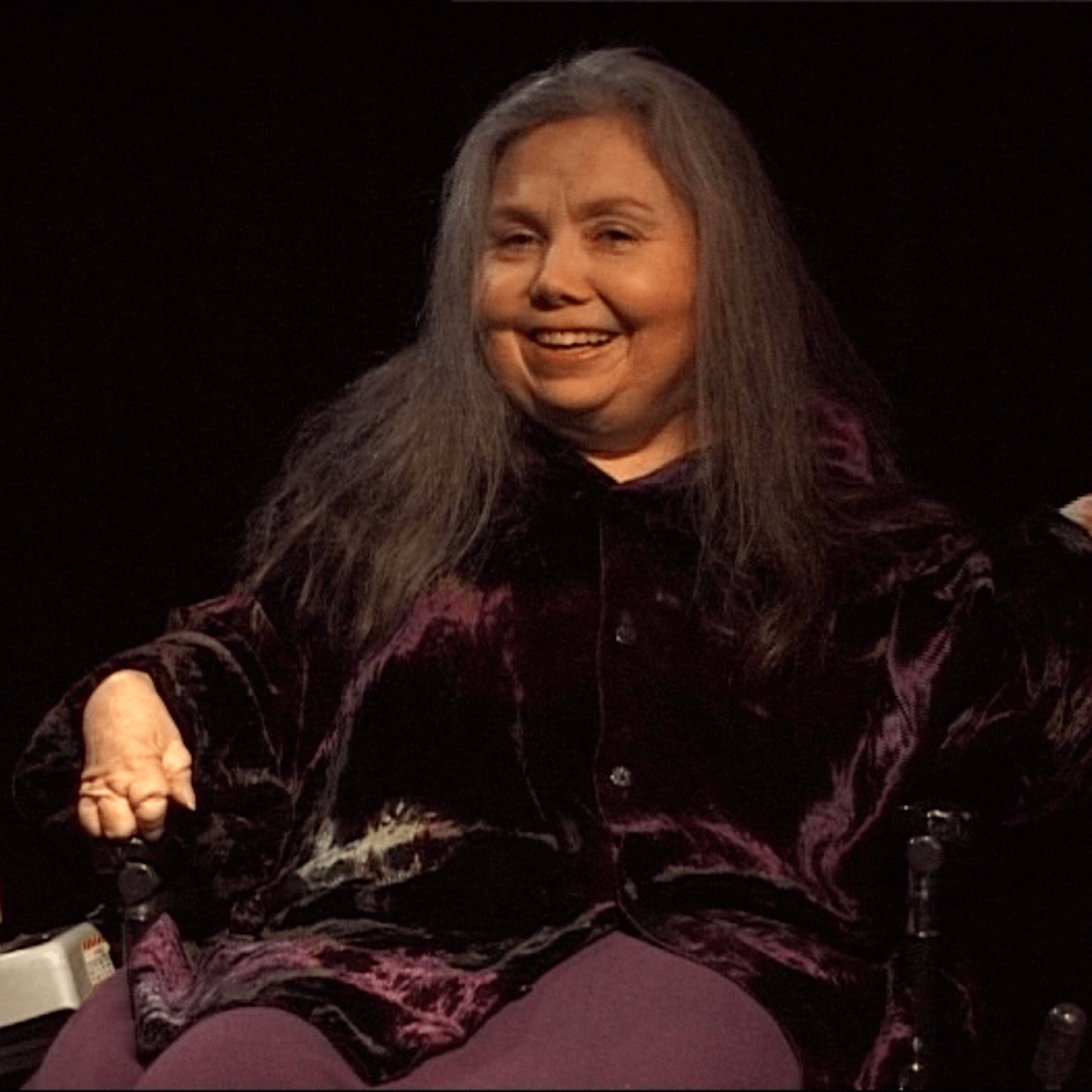 Une femme aux longs cheveux gris est assise dans un fauteuil roulant, vêtue d'un haut en velours foncé, et sourit sur un fond sombre.