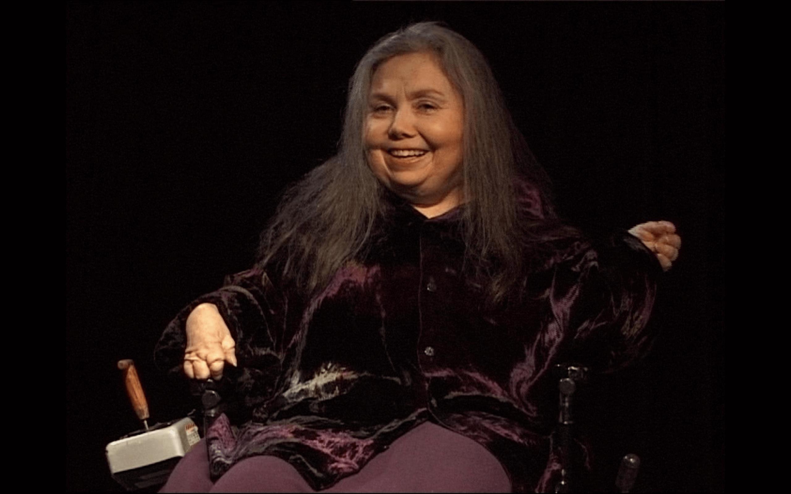 Une femme aux longs cheveux gris, en fauteuil roulant électrique, vêtue d'un haut en velours foncé et souriante, est assise sur un fond noir.