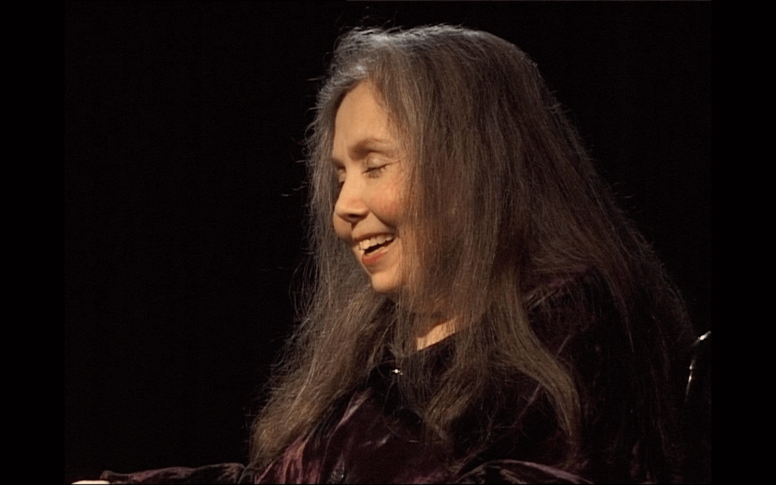 Une femme aux longs cheveux gris, vêtue d'un haut sombre, est assise et sourit, les yeux fermés, sur un fond noir.