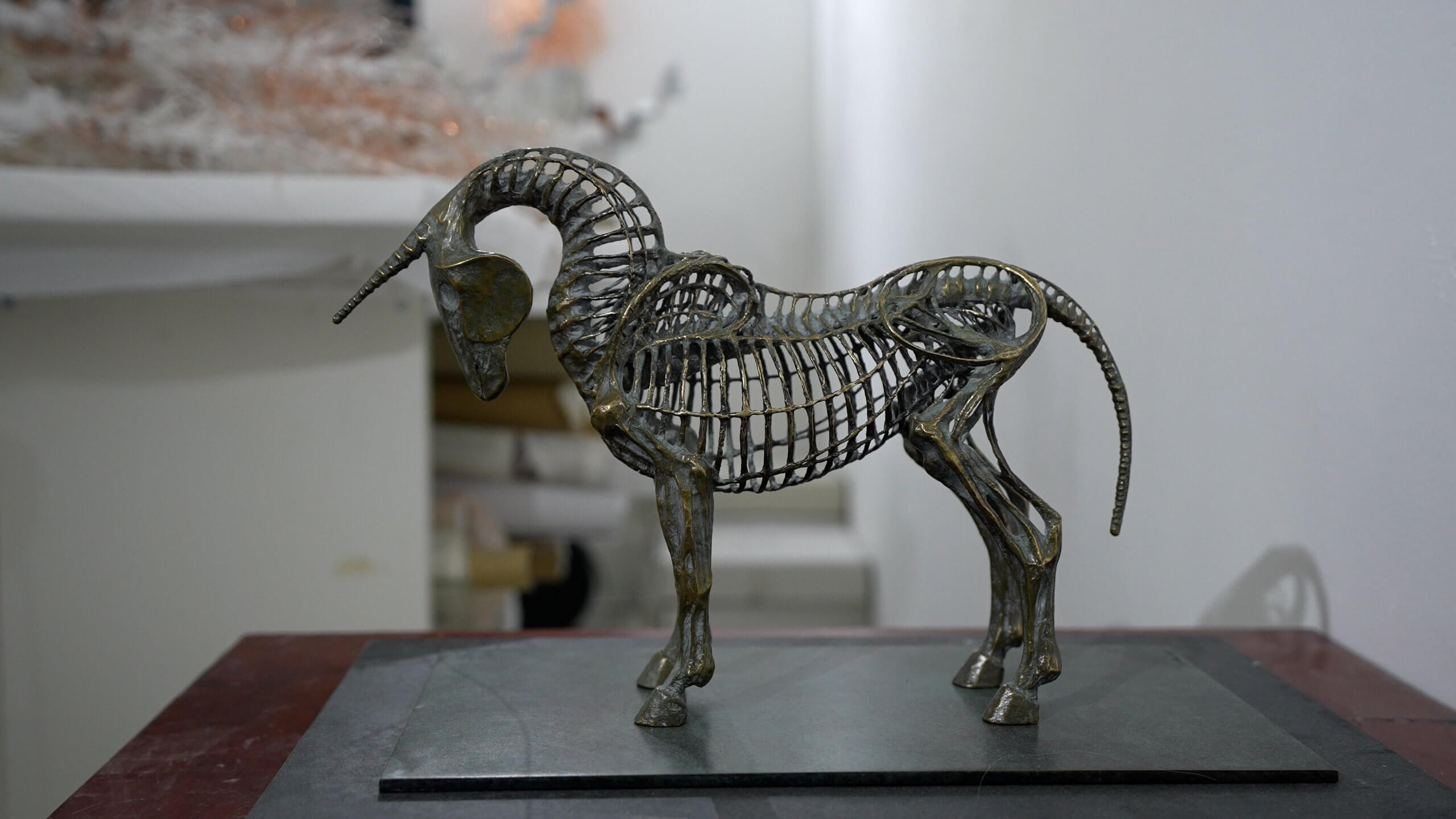 Une sculpture métallique représentant un squelette d'animal avec une corne incurvée est posée sur une surface plane sur un fond uni.