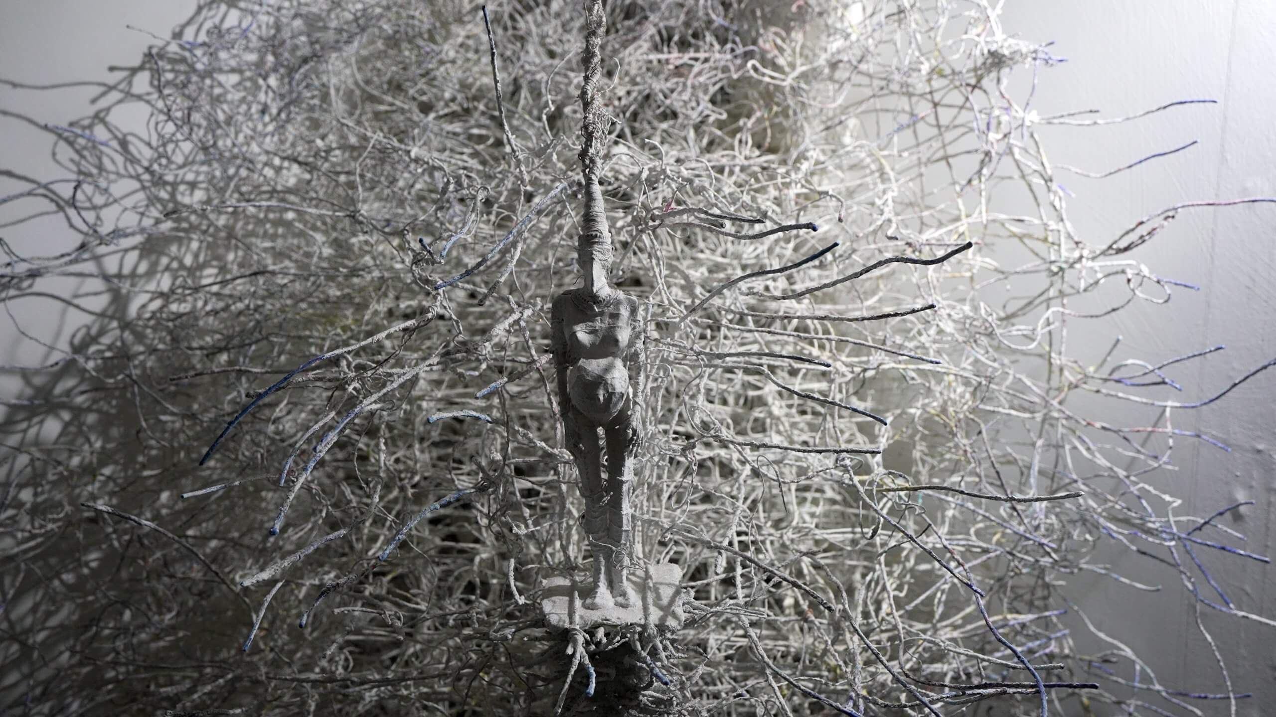 Une silhouette humaine grise se tient au milieu d'un enchevêtrement de fils ou de bâtons blancs en forme de branches, contre un mur ordinaire.