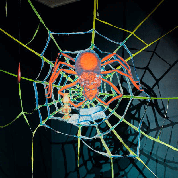 Une araignée aux couleurs vives et au visage humain est assise sur une toile, à côté d'un livre ouvert et d'un chandelier, sur un fond sombre.