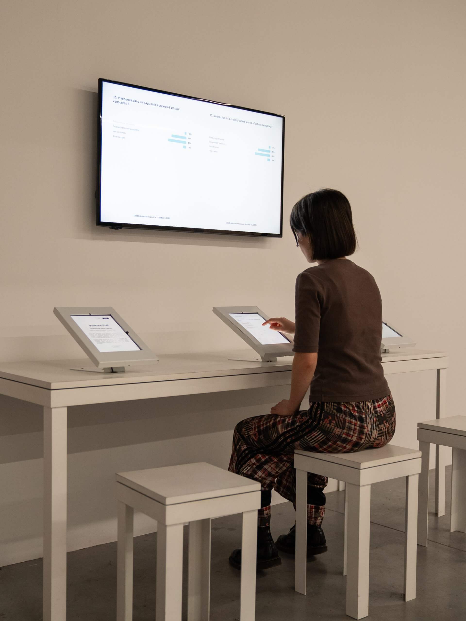 A person sits at a white desk using a touchscreen device, with two other tablets on the desk and a wall-mounted screen displaying bar charts in front of them.