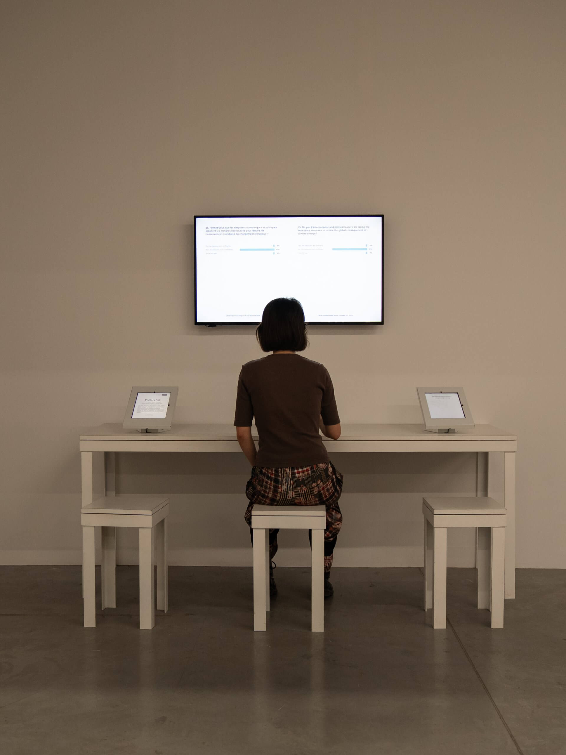 Une personne est assise à un bureau blanc avec deux tablettes, face à un écran mural affichant du texte et des graphiques dans une pièce minimaliste.
