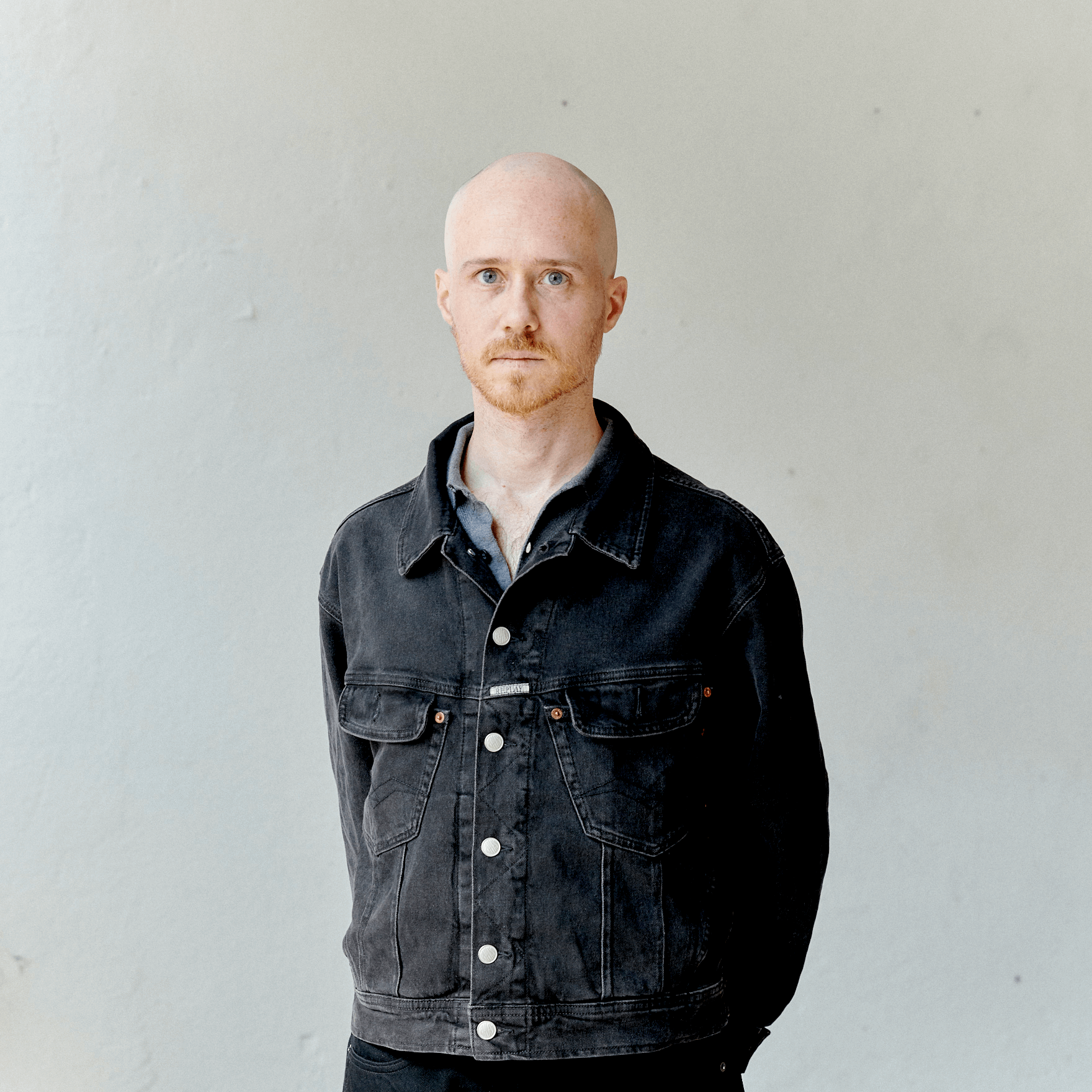 Un homme chauve à la barbe rousse, vêtu d'une veste en jean noire, se tient sur un fond uni de couleur claire et regarde directement la caméra.