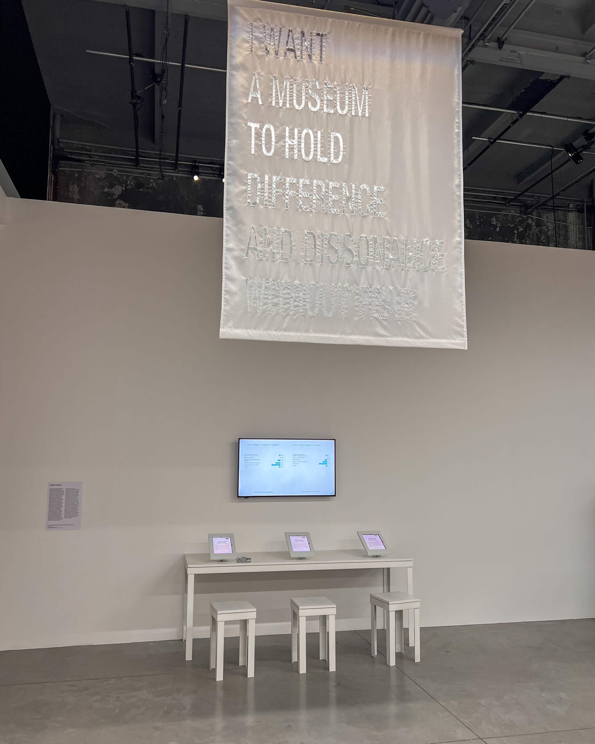 Une bannière blanche avec un texte brodé est suspendue au-dessus d'un présentoir contenant trois tablettes sur une table blanche, sous un moniteur mural dans une galerie contemporaine.