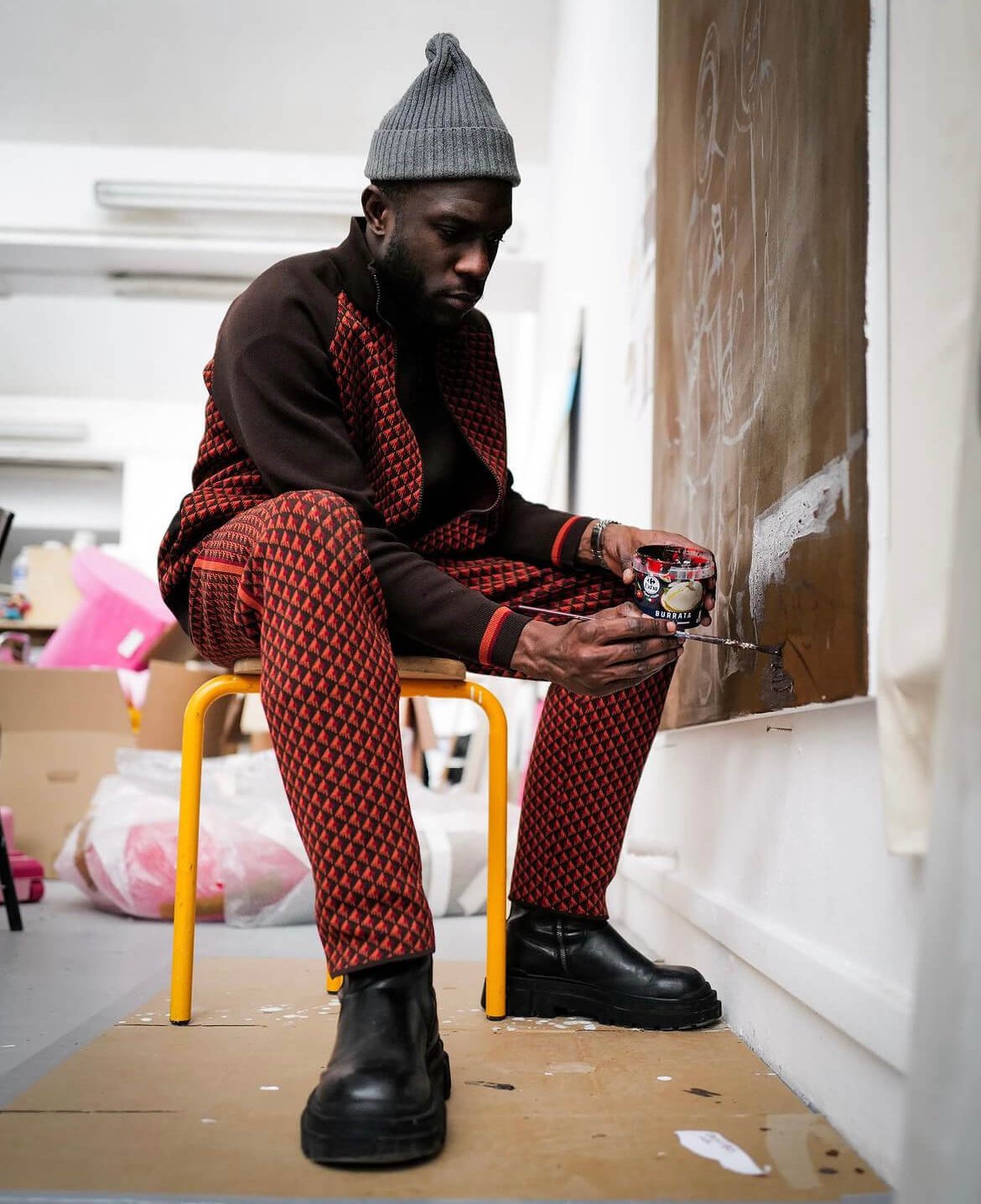 Une personne vêtue d'une tenue à motifs rouges et noirs est assise sur un tabouret jaune, peignant une toile brune dans un atelier d'art.