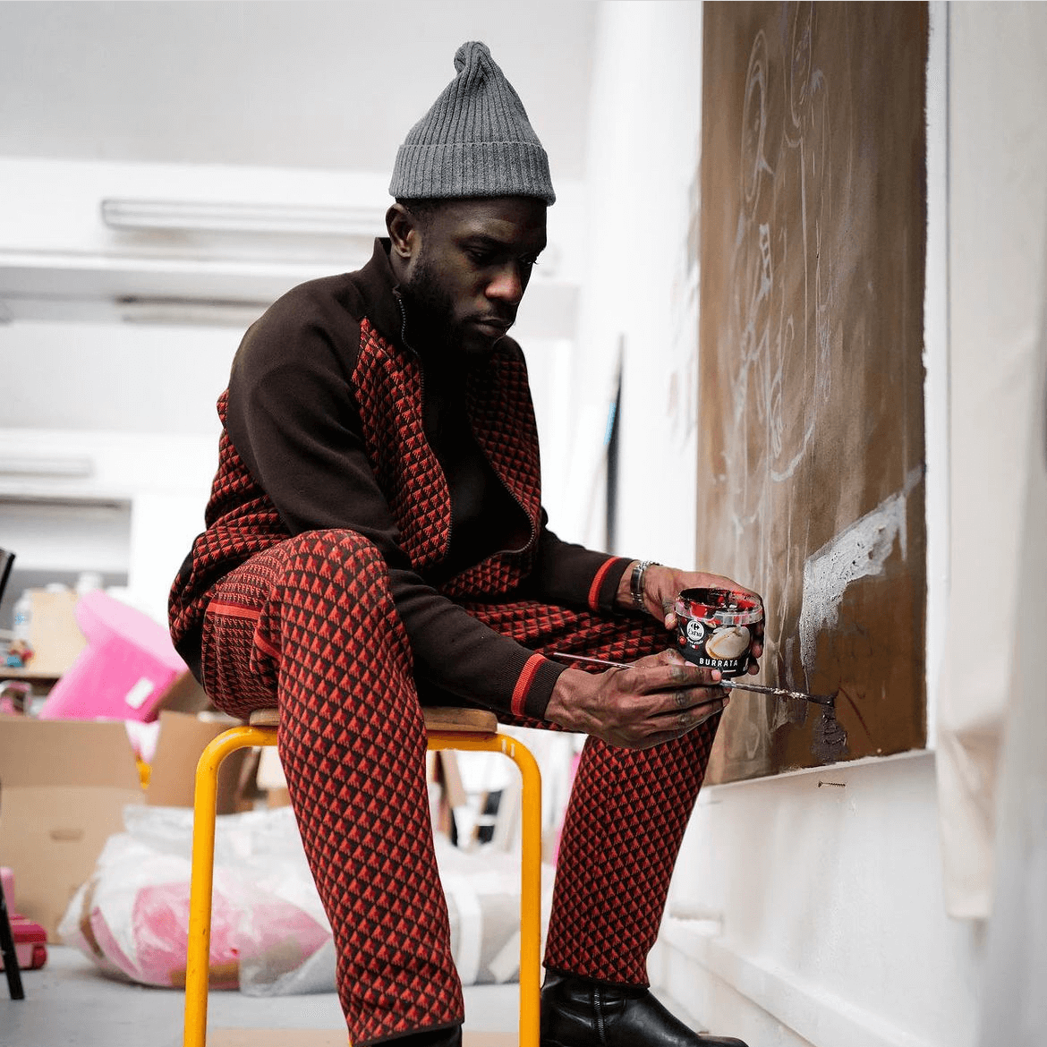 Un homme vêtu d'un ensemble à motifs rouges et noirs et d'un bonnet gris est assis sur un tabouret jaune, tenant une tasse et regardant une grande toile brune dans un atelier d'art.