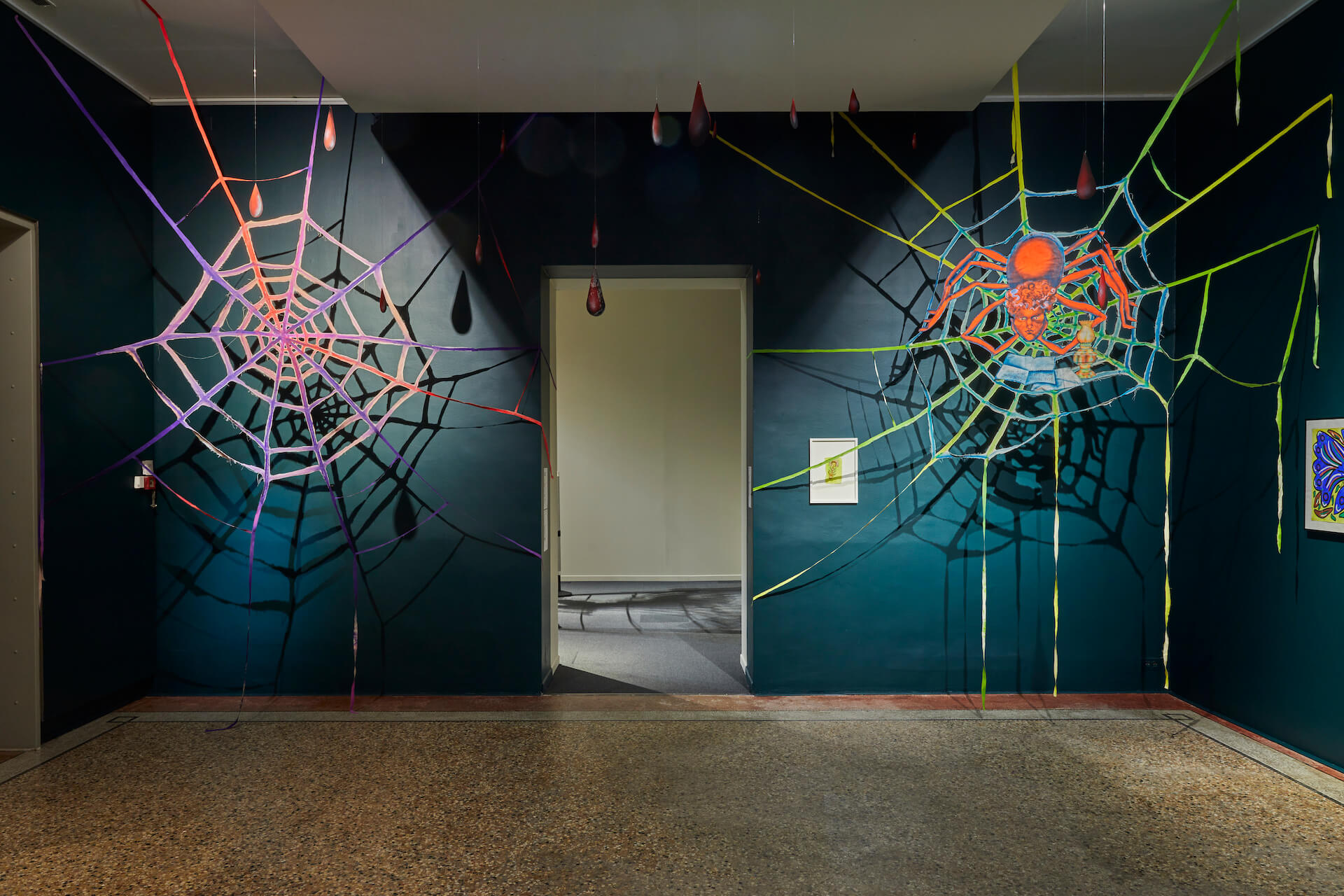 Deux toiles d'araignée artificielles colorées avec des gouttelettes suspendues sont exposées sur les murs sombres d'une galerie d'art ; une porte les sépare.