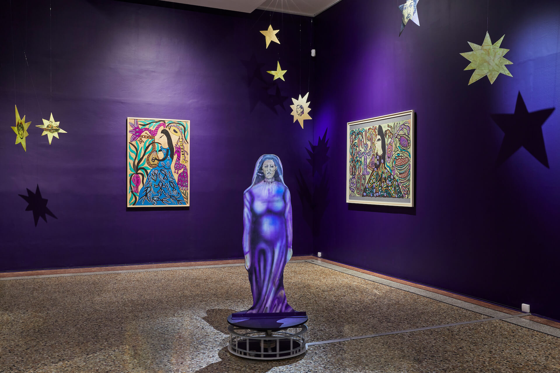 Une galerie d'art présente des murs violets, deux tableaux colorés, des décorations étoilées suspendues et un personnage violet grandeur nature au centre de la pièce.