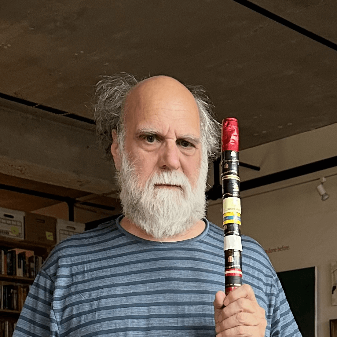 Un homme âgé à la barbe blanche et à la chemise rayée tient un bâton entouré de ruban adhésif de différentes couleurs, debout à l'intérieur, avec des étagères et des livres en arrière-plan.