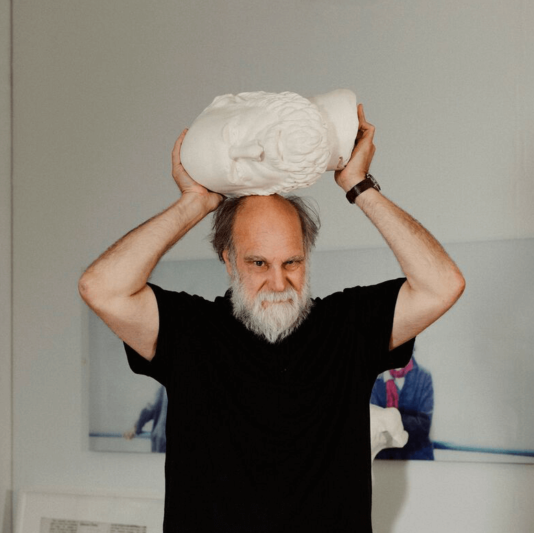 Un homme barbu et vêtu d'une chemise noire tient une tête sculptée blanche à l'envers sur sa propre tête, debout à l'intérieur.