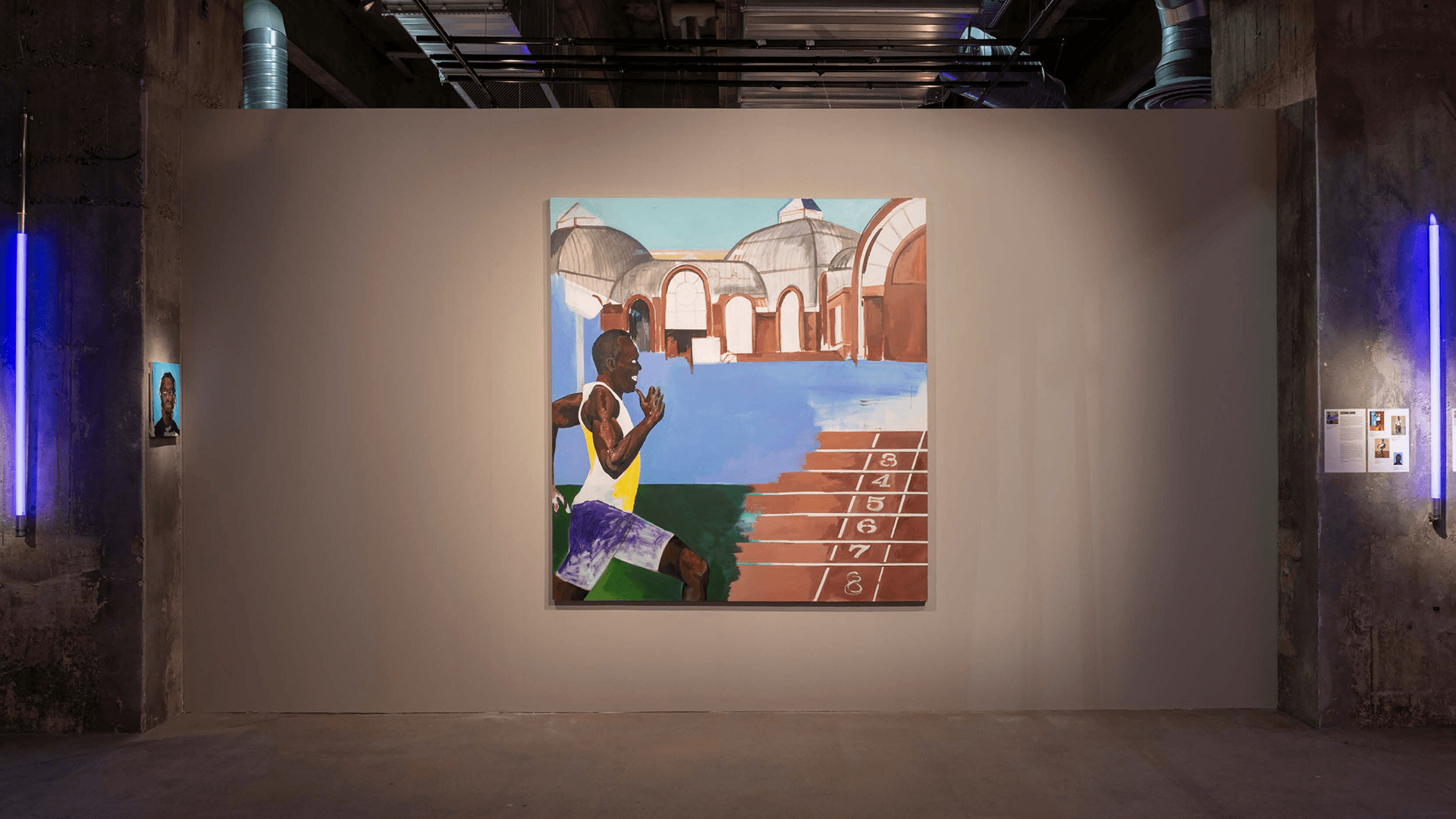 Une grande peinture représentant deux coureurs sur la ligne d'arrivée est exposée sur un mur gris d'une galerie industrielle, flanquée de néons bleus et d'œuvres d'art plus petites.