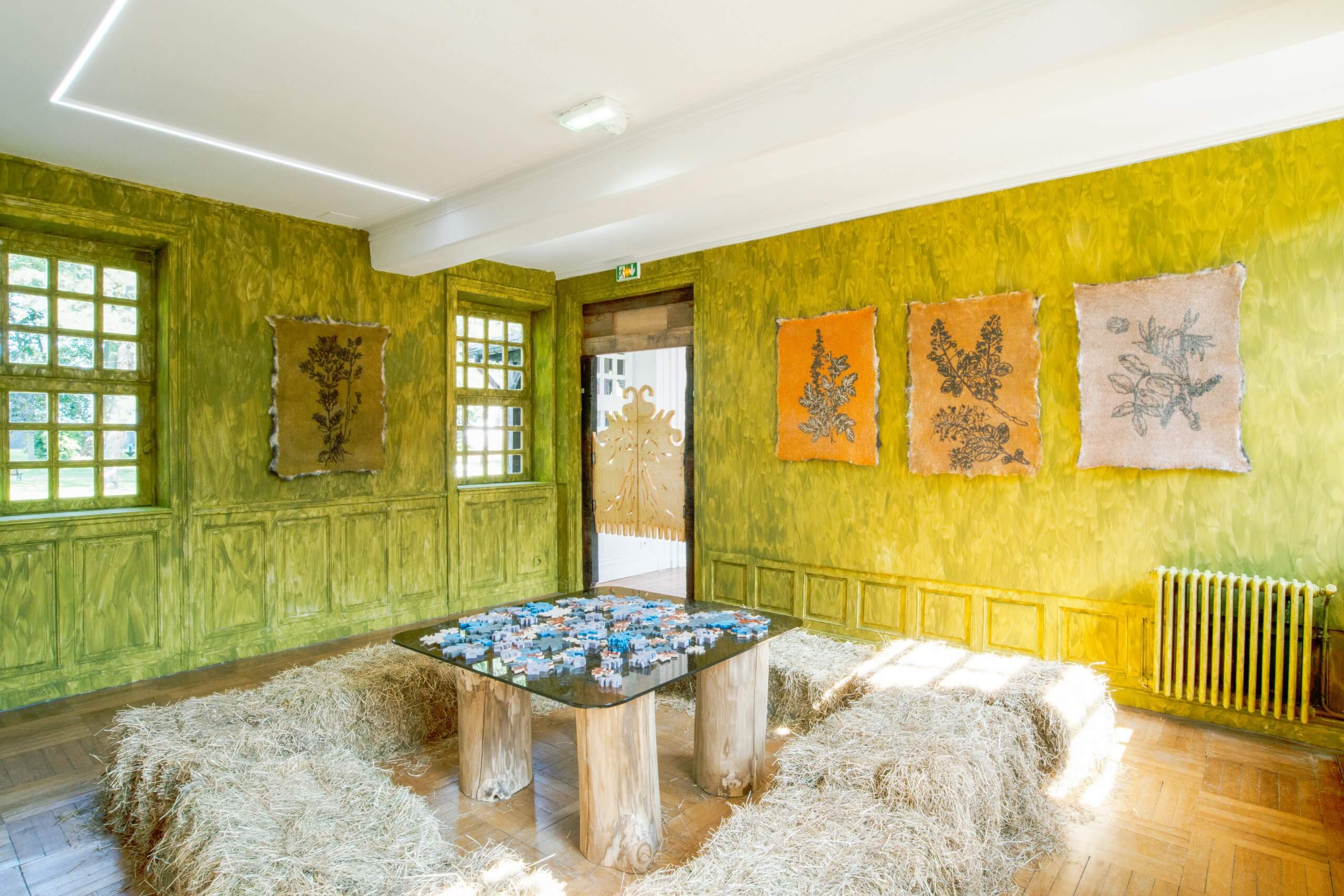 Une pièce lumineuse aux murs jaune verdâtre, des œuvres d'art botanique exposées, une table avec un puzzle et des bottes de paille disposées en guise de sièges autour de la table.