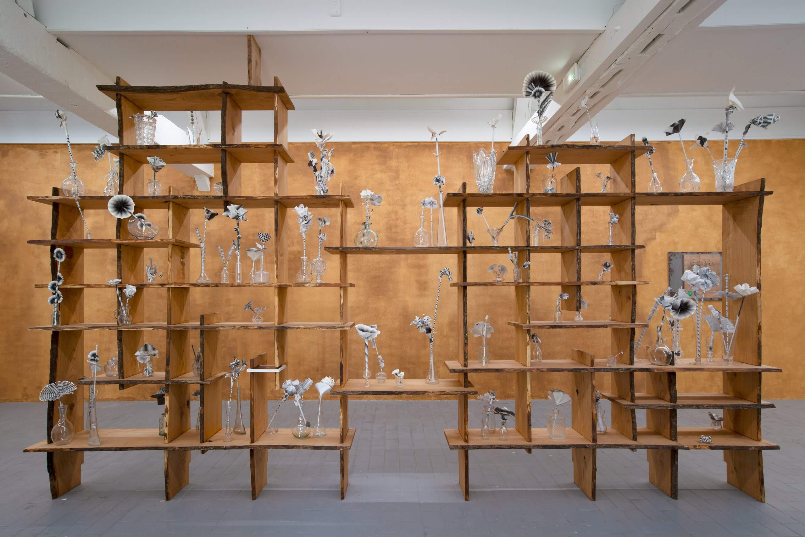 Une grande étagère en bois présente de nombreux vases en verre délicats avec des fleurs en verre complexes, sur un mur brun chaud dans une galerie.