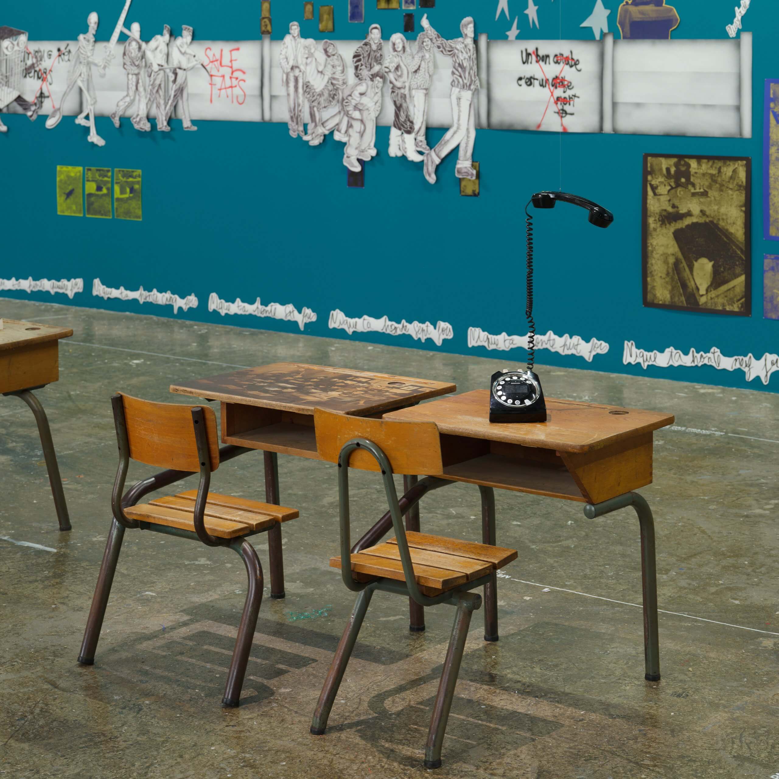 Two wooden desks and chairs sit on a concrete floor; an old rotary phone with a detached receiver rests on one desk. Colorful wall art and graffiti are visible in the background.