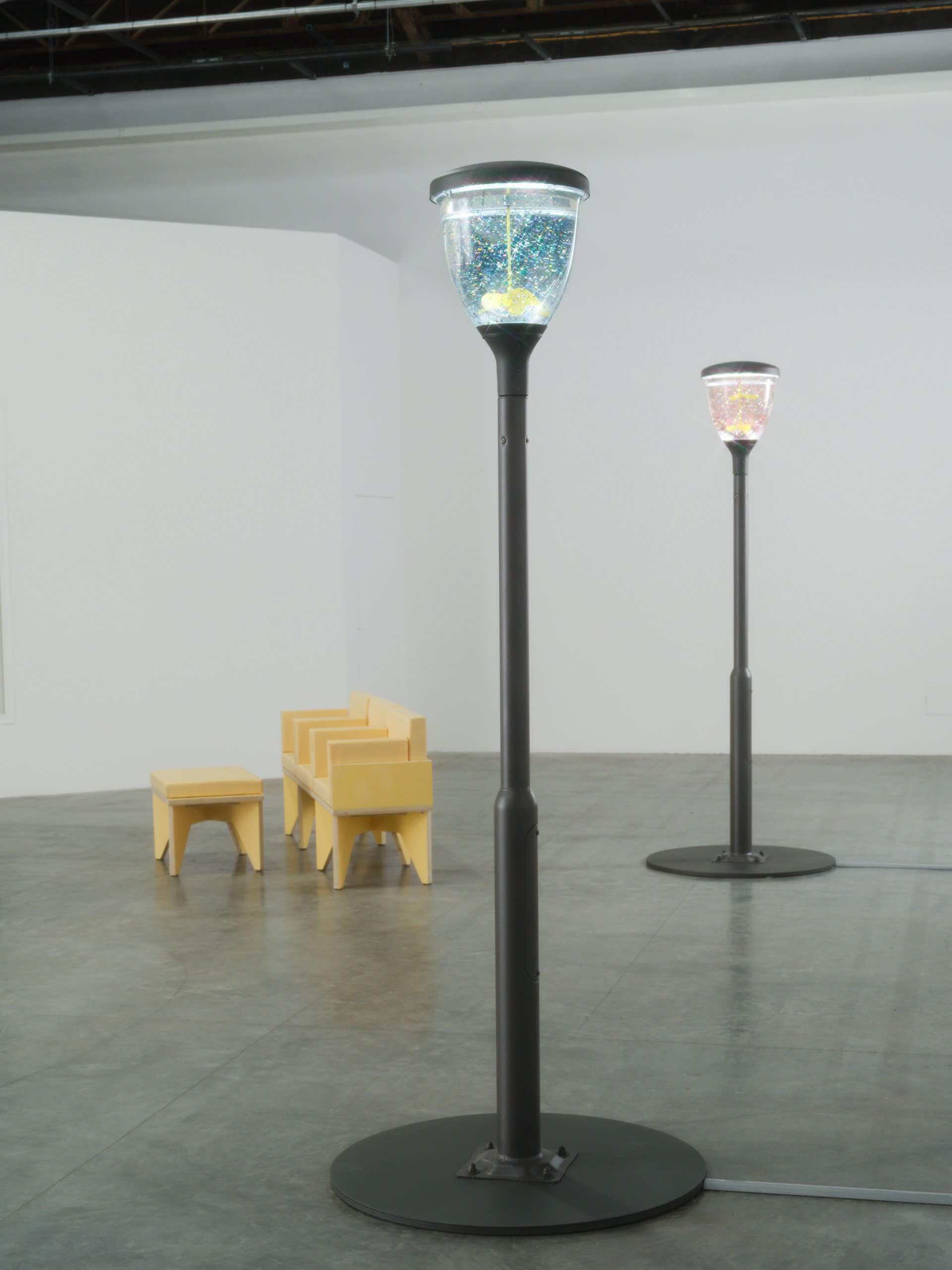 Deux lampadaires noirs avec des dessus en verre étincelants se dressent sur un sol en béton gris dans une pièce minimaliste avec plusieurs chaises et tabourets jaune pâle à l'arrière-plan.