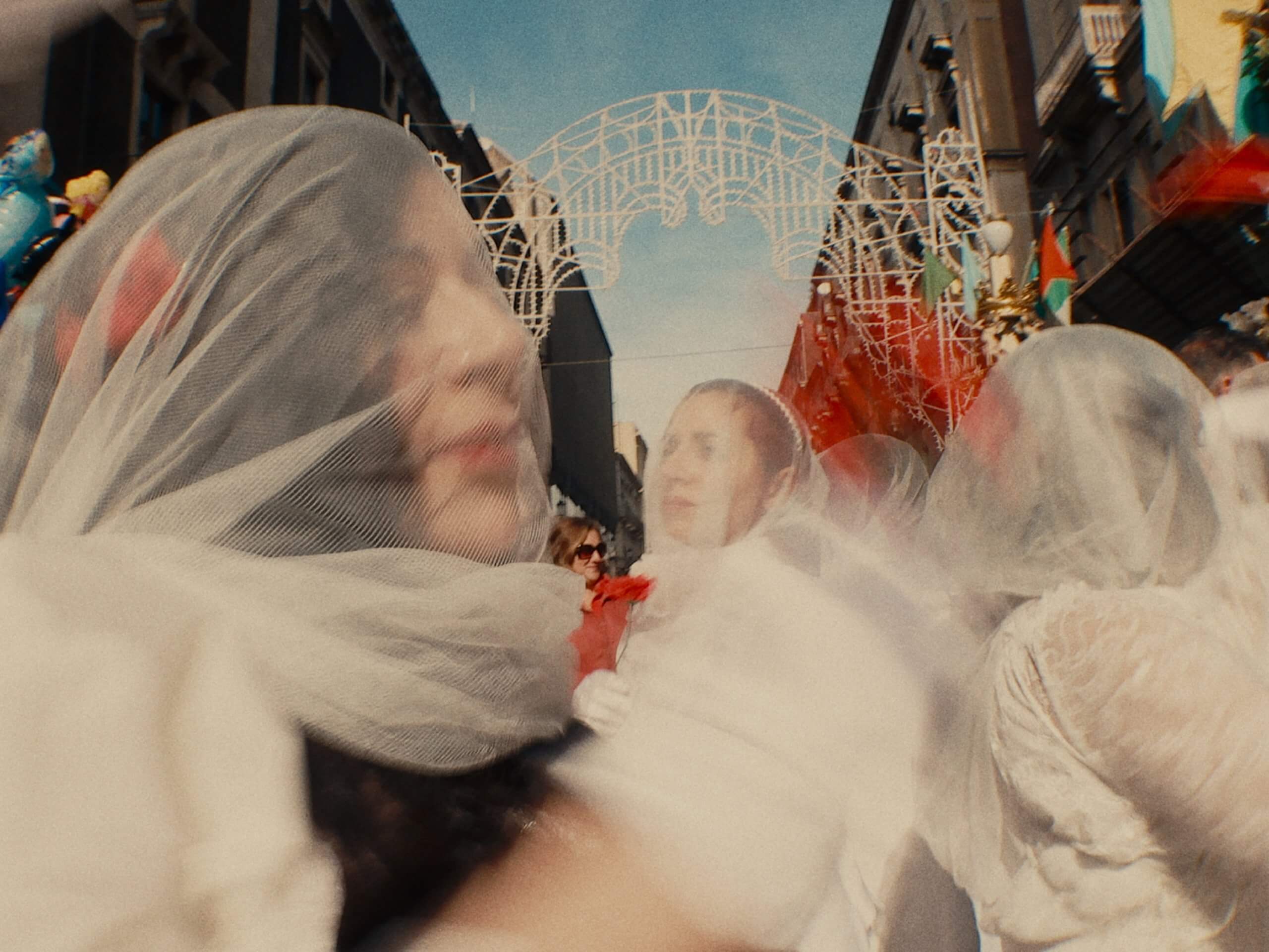 Des personnes vêtues de blanc et voilées participent à un défilé de rue sous des arches décoratives, le flou suggérant le mouvement.