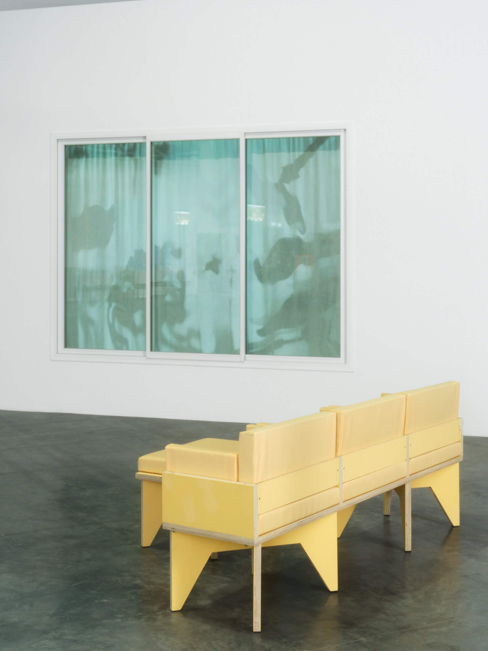 Un banc jaune minimaliste est posé sur un sol en béton poli devant une grande fenêtre aux rideaux verdâtres, adossée à un mur blanc uni.