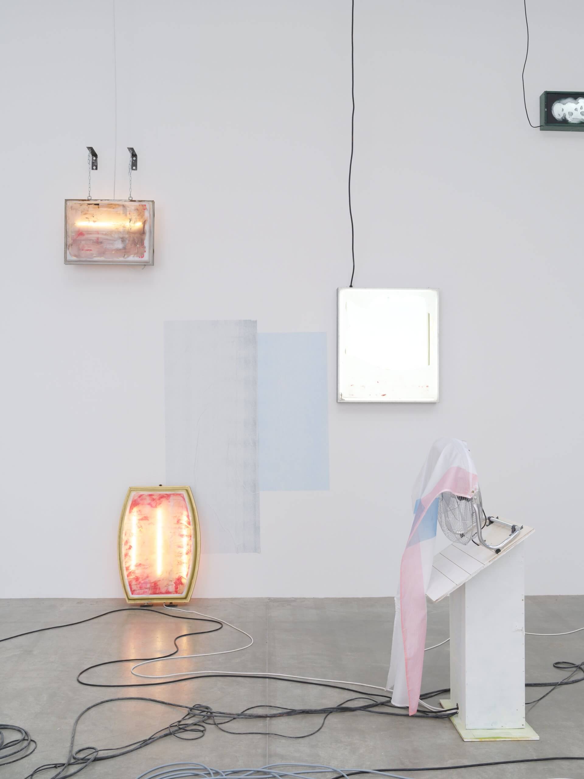 Installation artistique minimaliste comprenant des caissons lumineux sur un mur blanc, des tissus drapés, des câbles au sol et une petite plate-forme blanche avec du matériel électronique.