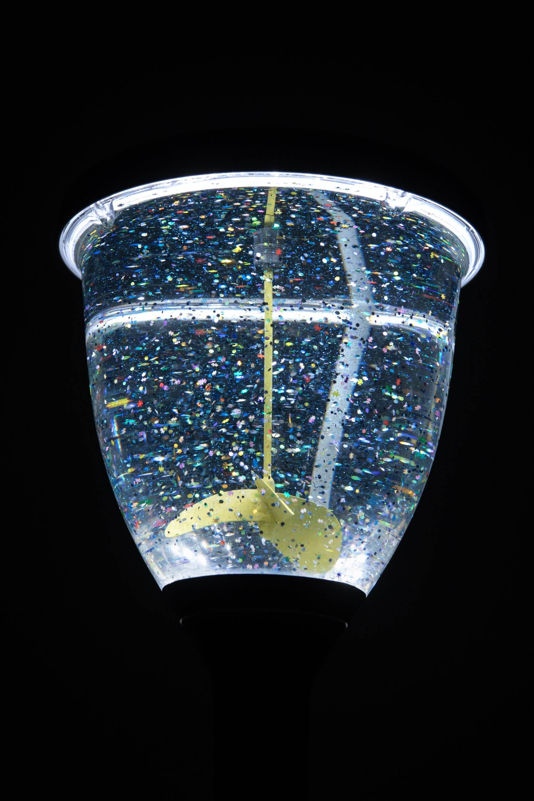 Gros plan sur une lampe remplie d'un liquide transparent et de paillettes en suspension, éclairée par le haut, avec une palette dorée à l'intérieur.