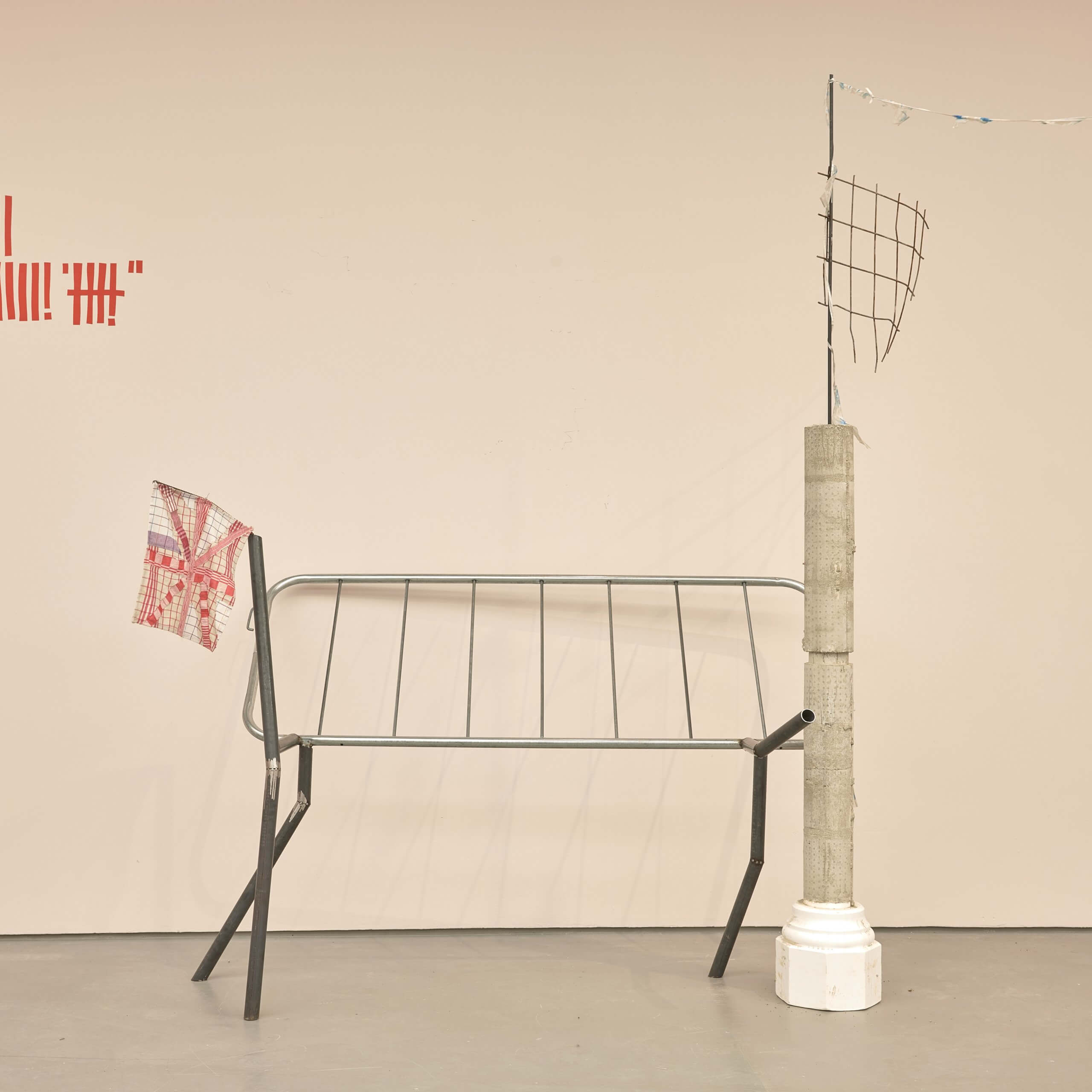 Une sculpture minimaliste composée d'une barrière métallique, d'un pilier en béton, d'un treillis métallique et d'un sac en plastique, disposée contre un mur beige uni avec des marques rouges.