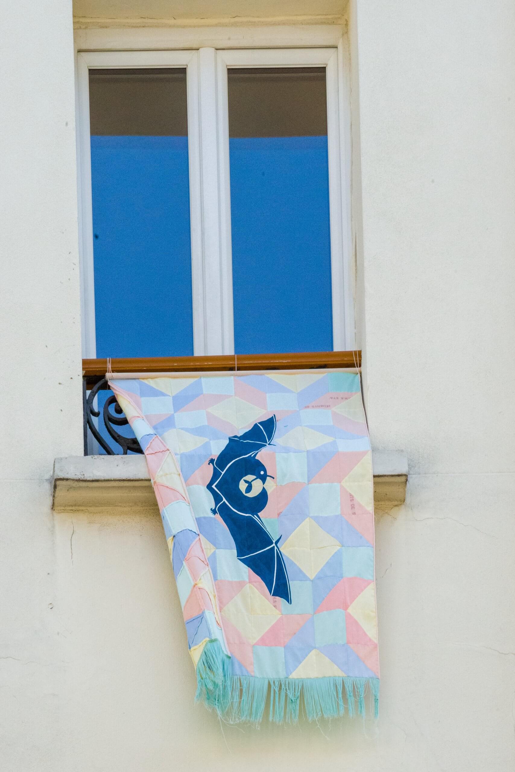 Une couverture à motifs d'oiseaux géométriques est suspendue sur le rebord d'une fenêtre d'un bâtiment blanc.