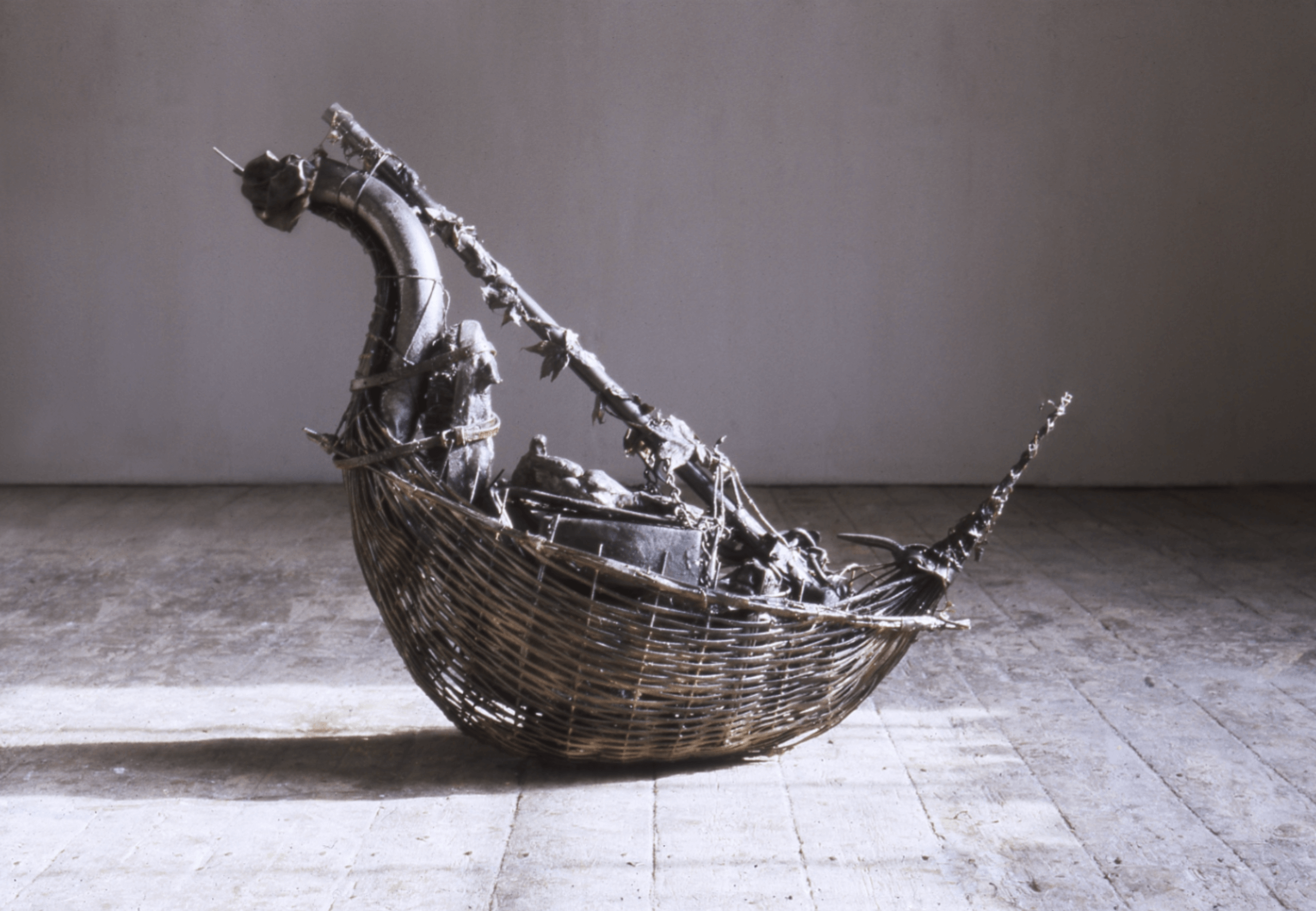Un panier tressé en forme de bateau, rempli de divers objets sombres et sculpturaux et de branches, est posé sur un sol en bois contre un mur uni.