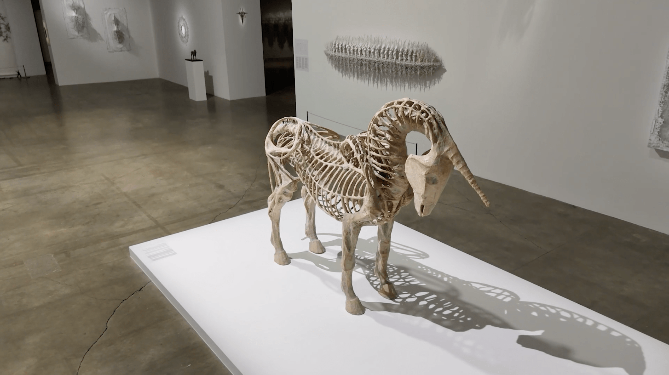 Une sculpture en bois ressemblant à un squelette d'animal, probablement une chèvre ou un bélier, est exposée sur une plate-forme blanche dans une galerie d'art moderne.