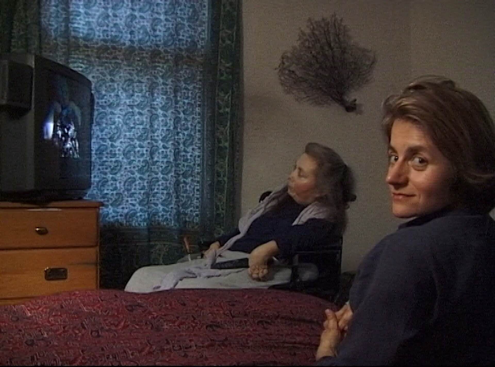 A woman in a wheelchair watches TV beside a window, while another woman sits on a bed in the foreground, looking toward the camera.