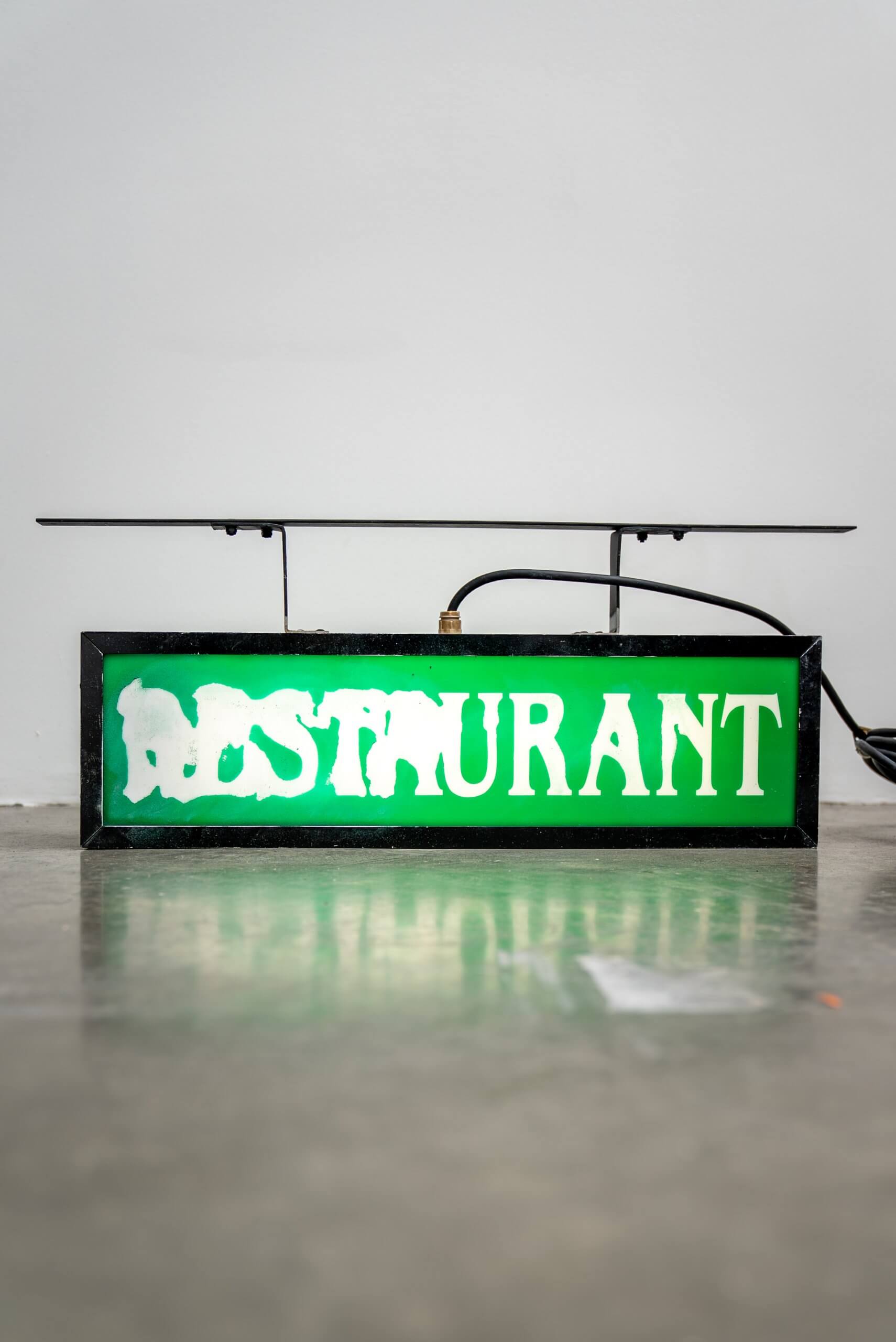 A rectangular green sign with the word RESTAURANT in white, with parts of the letters partially faded or obscured, sits on a gray floor against a plain white wall.