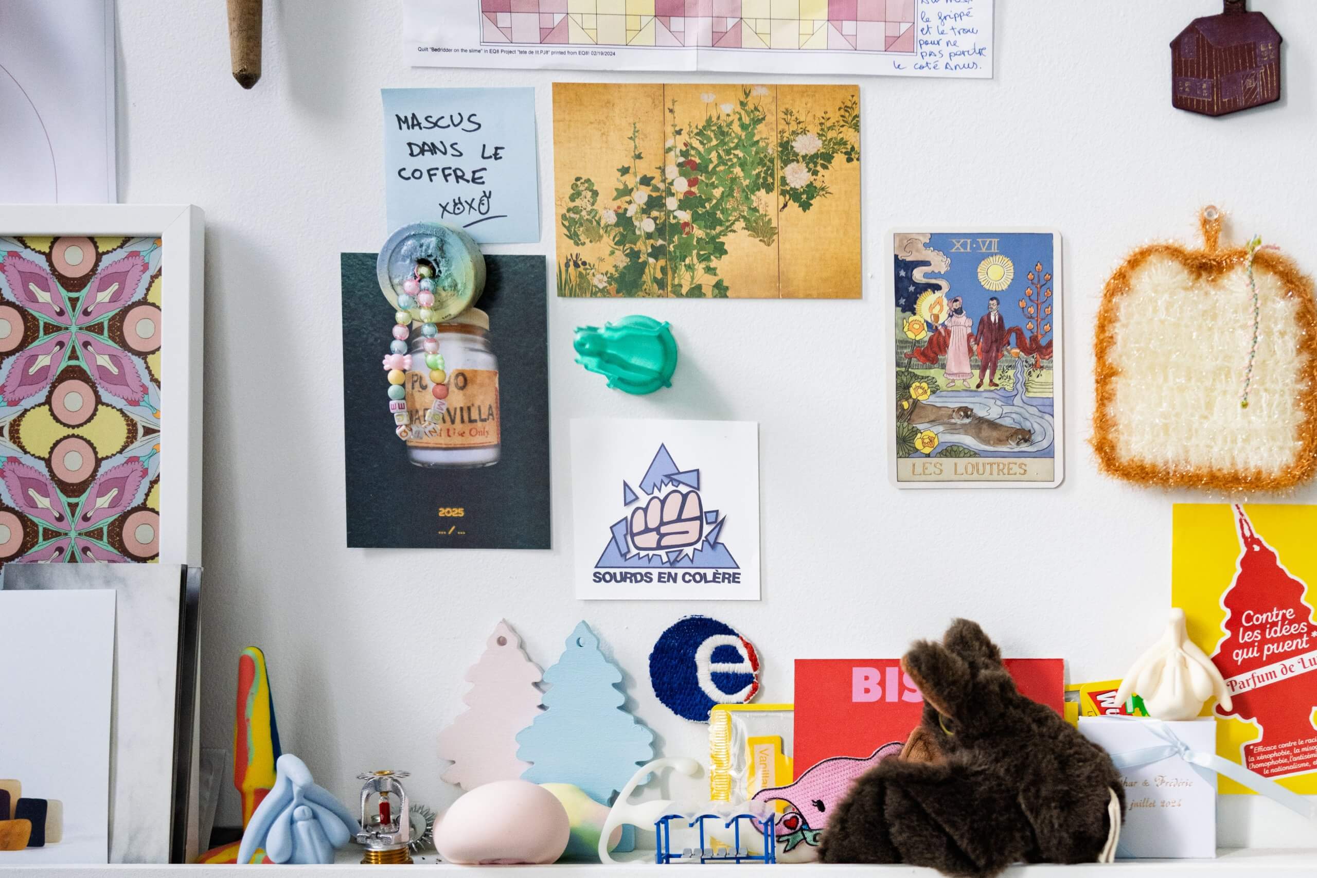 Un mur encombré de cartes postales, de dessins, d'un calendrier, de notes et de petits objets décoratifs au-dessus d'une étagère contenant des jouets, des cartes et des objets colorés.