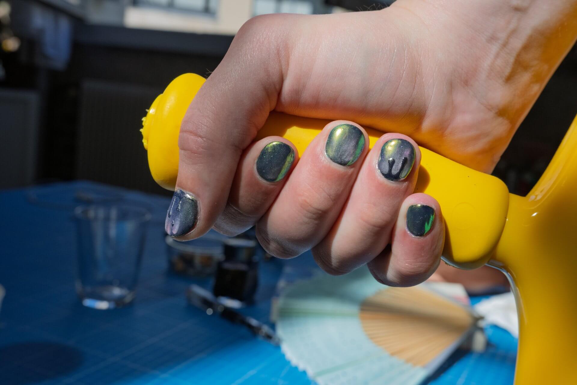 Une main recouverte de vernis à ongles vert métallique et violet saisit une poignée jaune ; la lumière du soleil fait ressortir les ongles et les éléments de l'arrière-plan sont flous.
