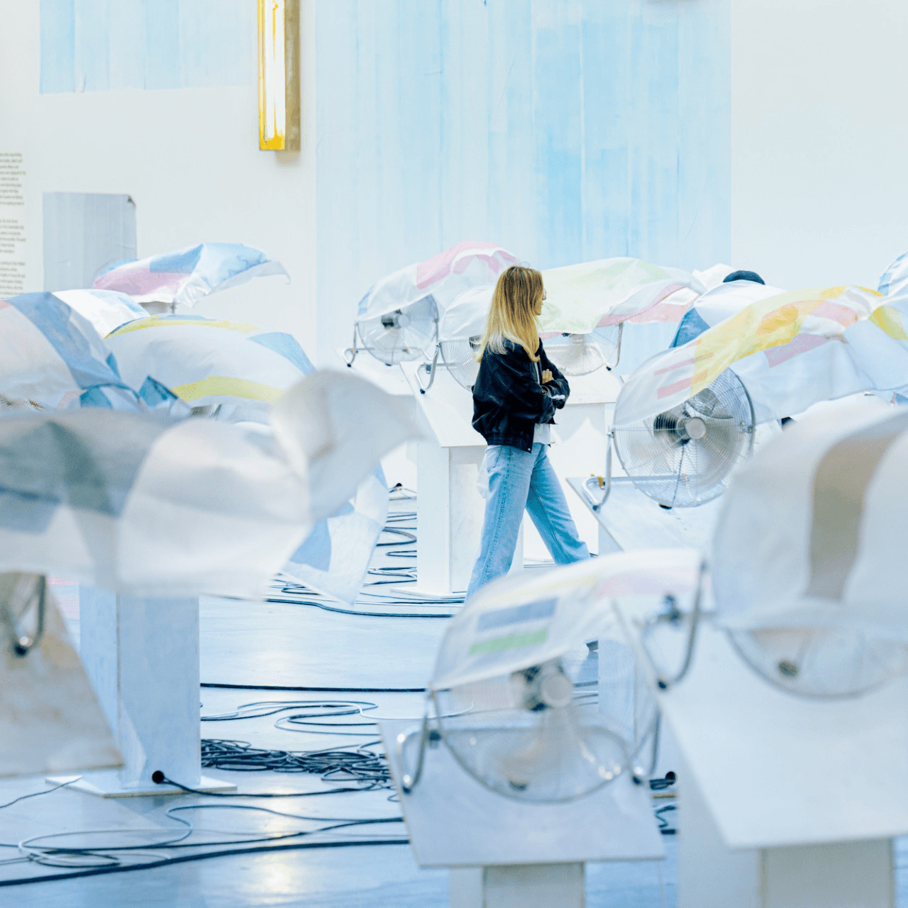 Une femme se tient au milieu d'installations artistiques représentant des ventilateurs recouverts d'un tissu translucide de couleur pastel dans une galerie très éclairée.