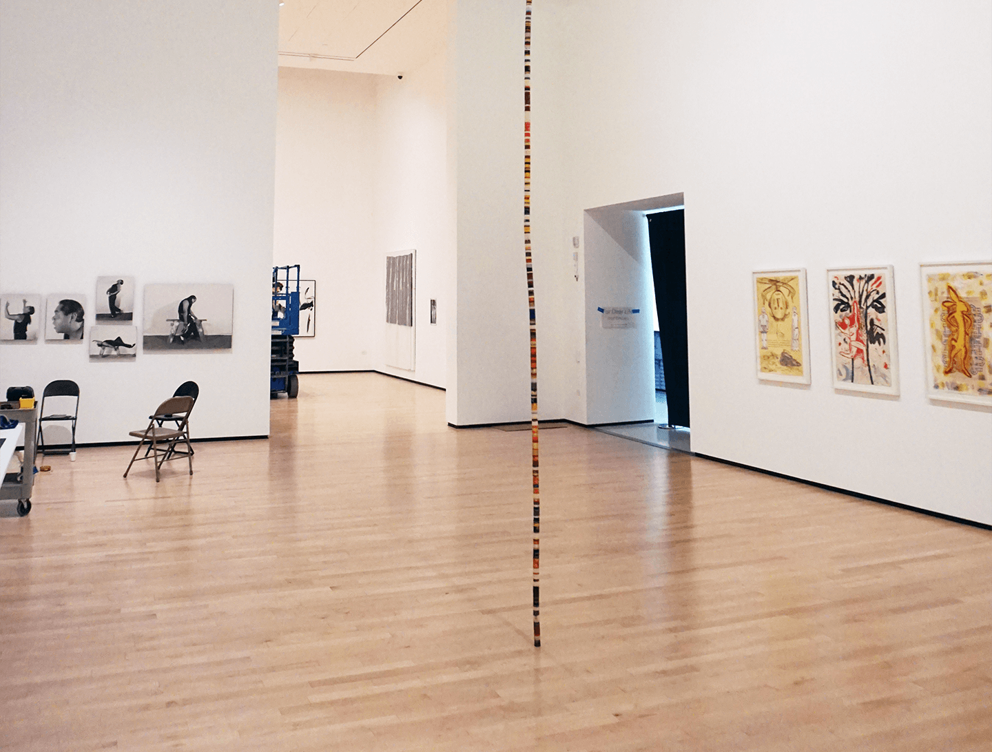 Une galerie d'art contemporain avec un sol en bois clair, diverses œuvres d'art encadrées sur des murs blancs, des chaises éparpillées et, au centre, une sculpture haute et fine faite de blocs colorés.