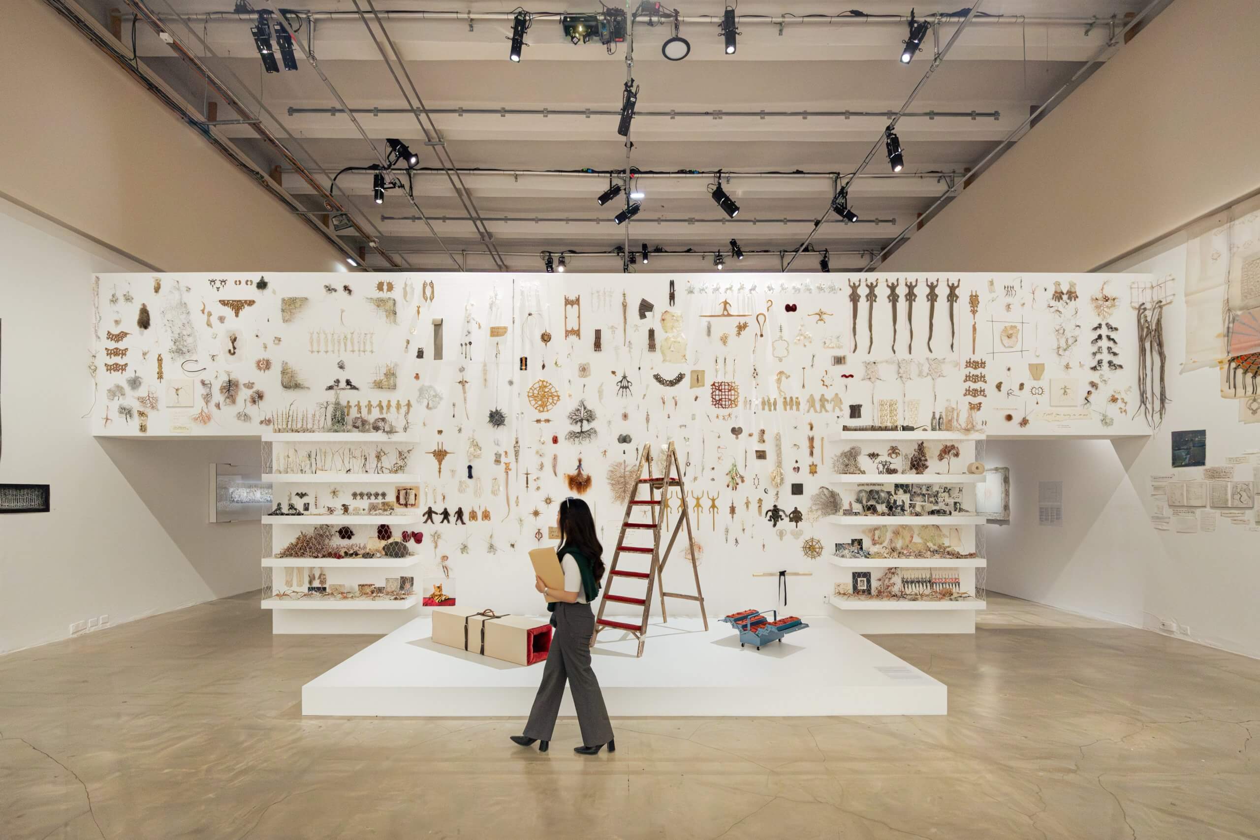 Une femme passe devant une installation artistique composée de divers objets et artefacts disposés sur un grand mur d'exposition blanc dans une galerie spacieuse.