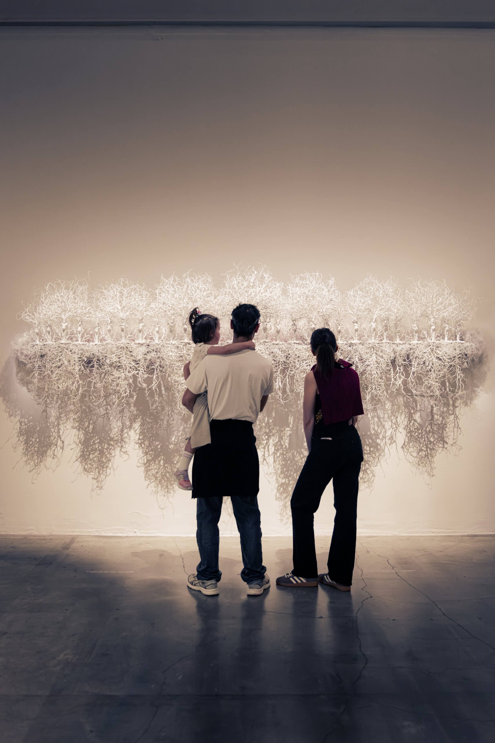 Trois personnes regardent une grande installation d'art abstrait blanc montée sur le mur d'une galerie, avec des textures complexes et enchevêtrées qui s'étendent vers l'extérieur.