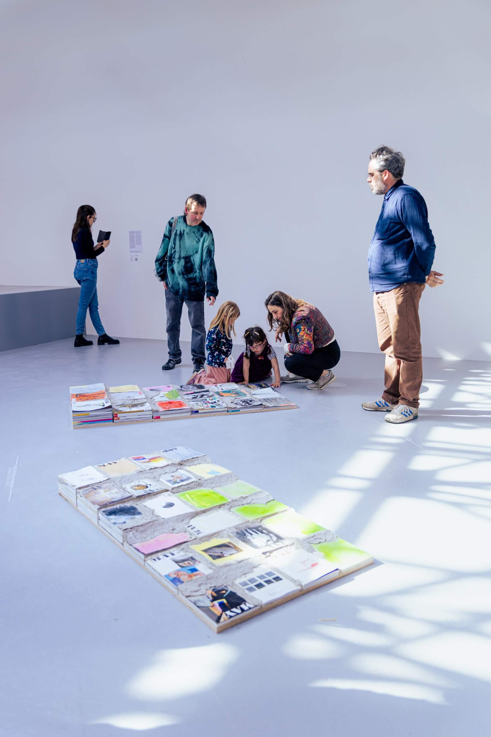 Several people in a brightly lit gallery view and interact with art pieces displayed on the floor; one person takes a photo, while others observe or kneel near the artwork.