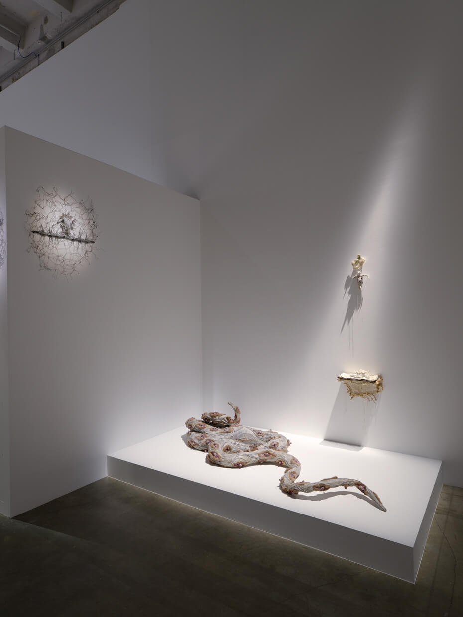Une galerie d'art minimaliste présente trois sculptures abstraites : une pièce murale enchevêtrée, une forme organique montée et une grande sculpture en forme de serpent sur une plate-forme blanche.
