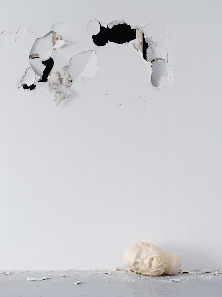 A damaged white wall with several large holes and a broken sculpture of a head lying on the floor beneath it.