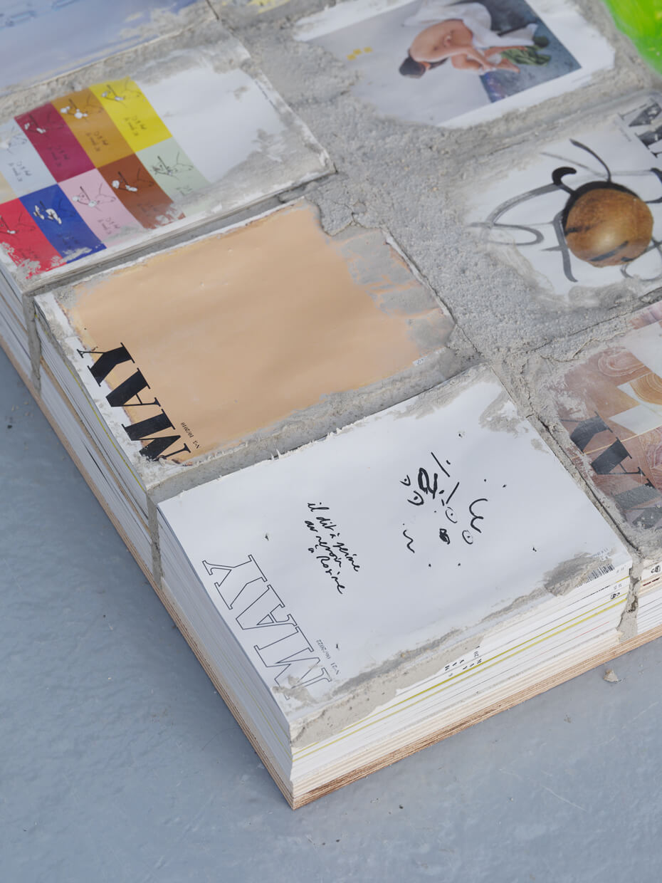 A stack of magazines, partially covered in concrete, shows various colorful and monochrome covers with text and images, laid on a smooth gray floor.
