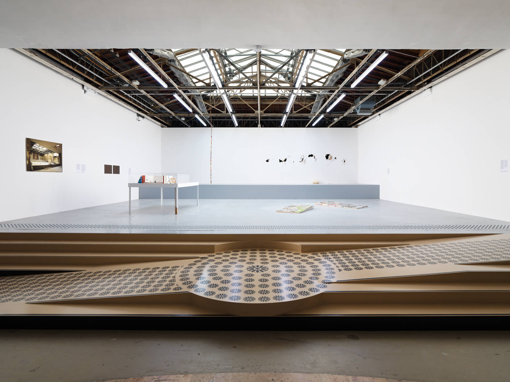 A modern art gallery with a skylight ceiling, white walls, various artworks displayed, and a beige patterned installation on the floor in the foreground.