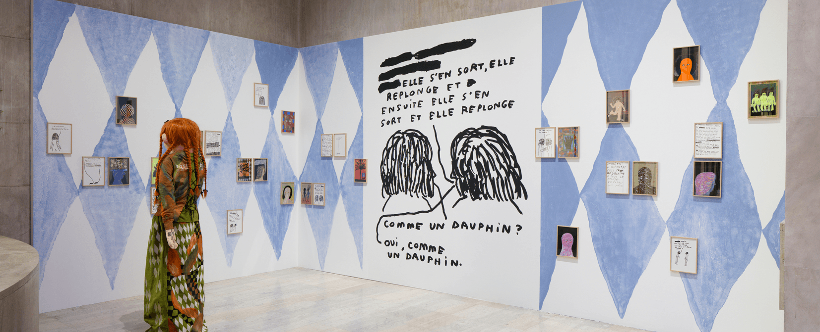 Une personne regarde une exposition d'art comprenant des tableaux encadrés et un grand mur illustré avec un texte en français et des croquis de deux personnages, sur un fond géométrique bleu et blanc.