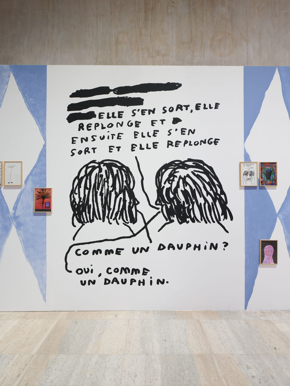 Une grande fresque murale représente deux personnages dessinés en noir avec un texte en français au-dessus et en dessous ; des œuvres d'art encadrées sont exposées de part et d'autre de la fresque.